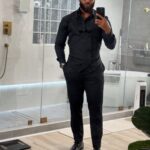 A man in an all-black button-down shirt, trousers, and dress shoes posing for a mirror selfie.
