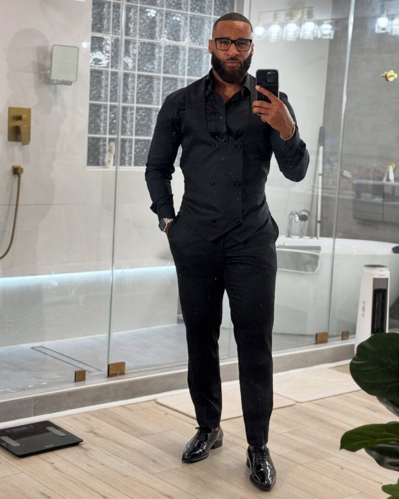 A man in an all-black button-down shirt, trousers, and dress shoes posing for a mirror selfie.