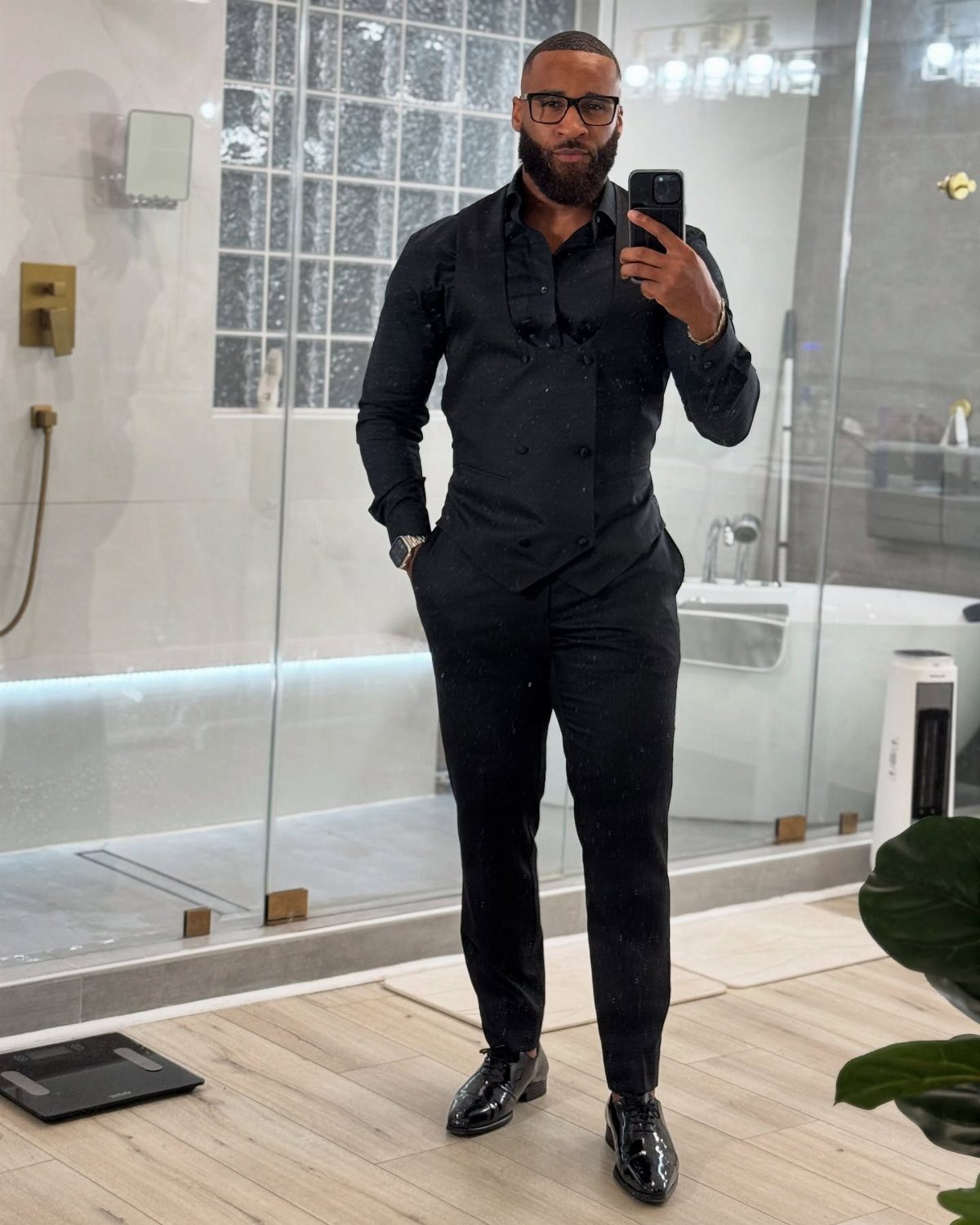 A man in an all-black button-down shirt, trousers, and dress shoes posing for a mirror selfie.