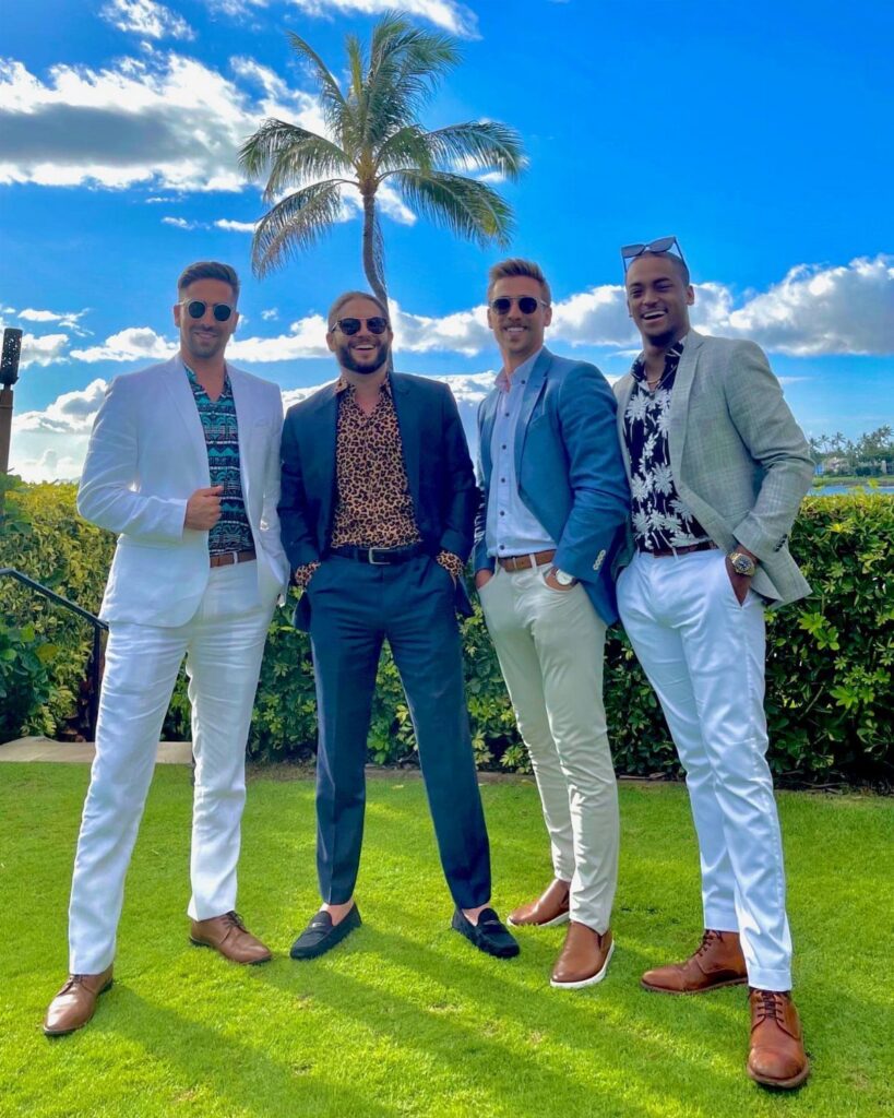 Four men in stylish summer suits posing on a sunny lawn with a palm tree.