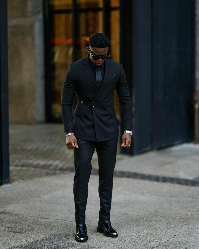 A man in a black double-breasted suit, black shirt, and tie posing outdoors.