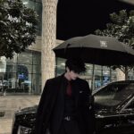 A person in a black suit and red tie standing under a black umbrella by a car.