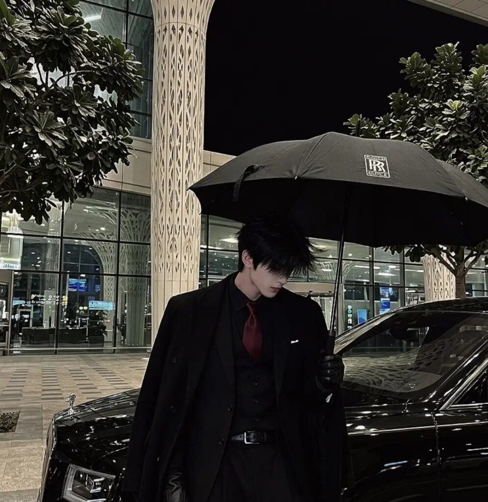 A person in a black suit and red tie standing under a black umbrella by a car.