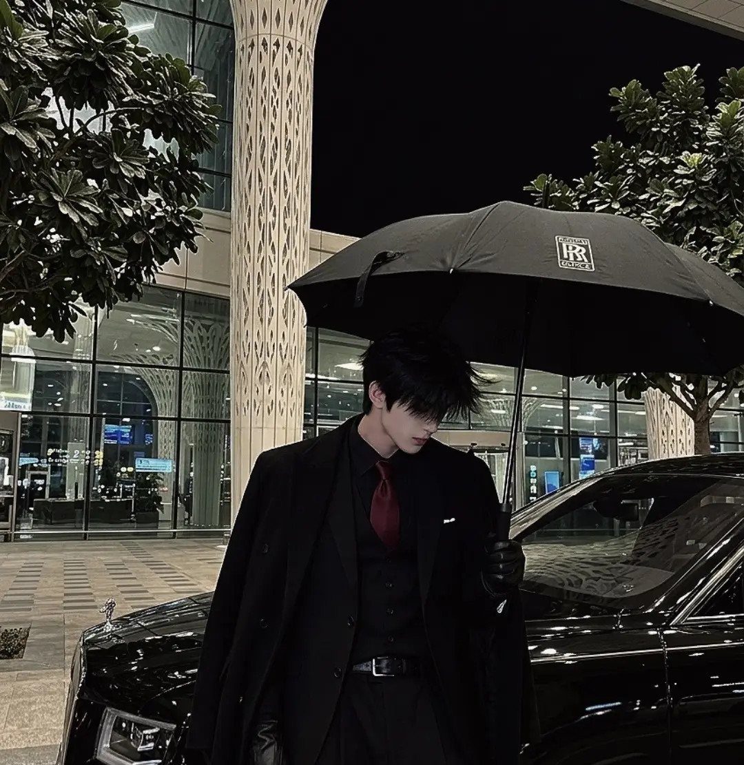 A person in a black suit and red tie standing under a black umbrella by a car.