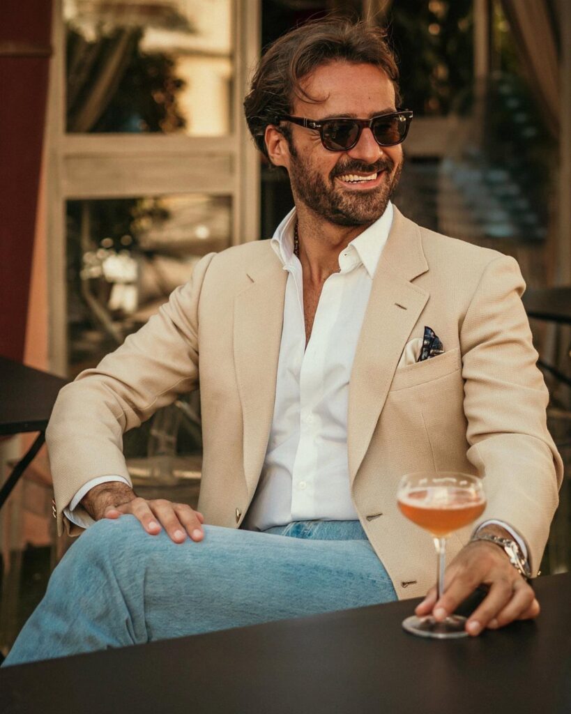 A man in a beige cotton blazer and light blue jeans sitting at a cafe table.