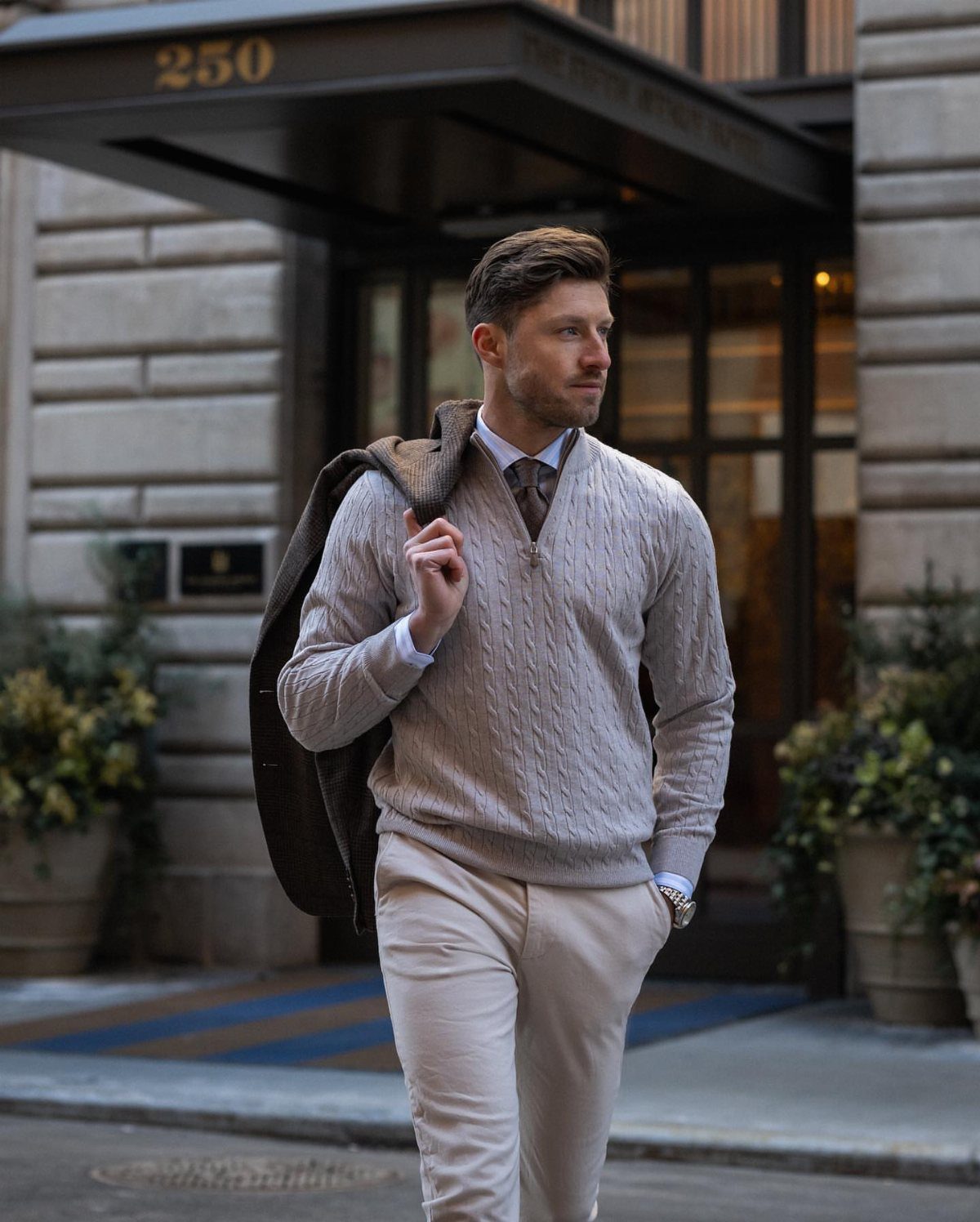 Man wearing a grey cable-knit sweater over a collared shirt and tie while walking outdoors.