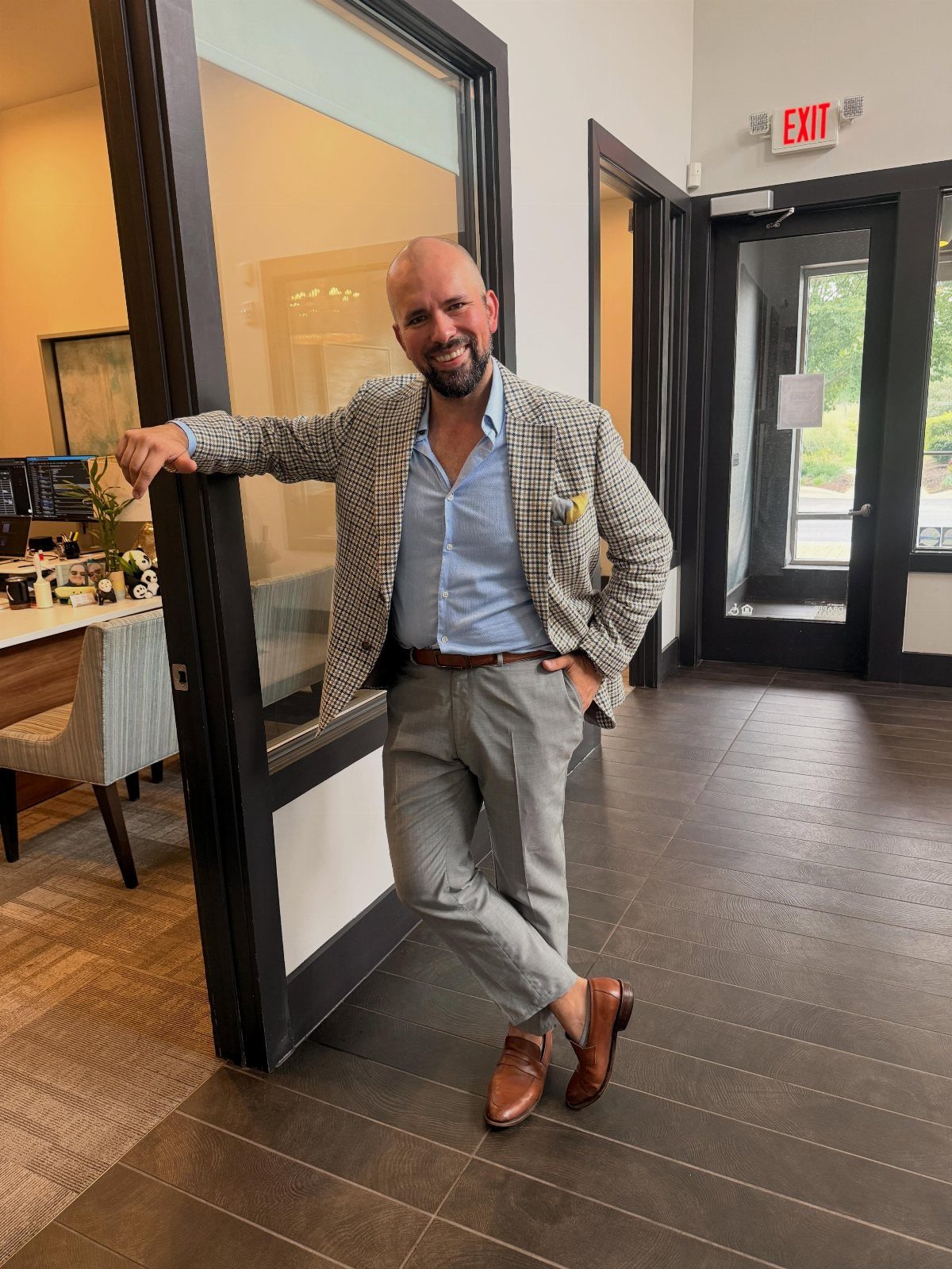 A man in a patterned blazer and grey trousers leaning against a glass door.
