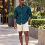 A man wearing a teal button-down shirt and cream shorts walking outdoors.