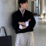 Man in a black knit sweater layered over a white shirt and beige trousers leaning against a pillar.