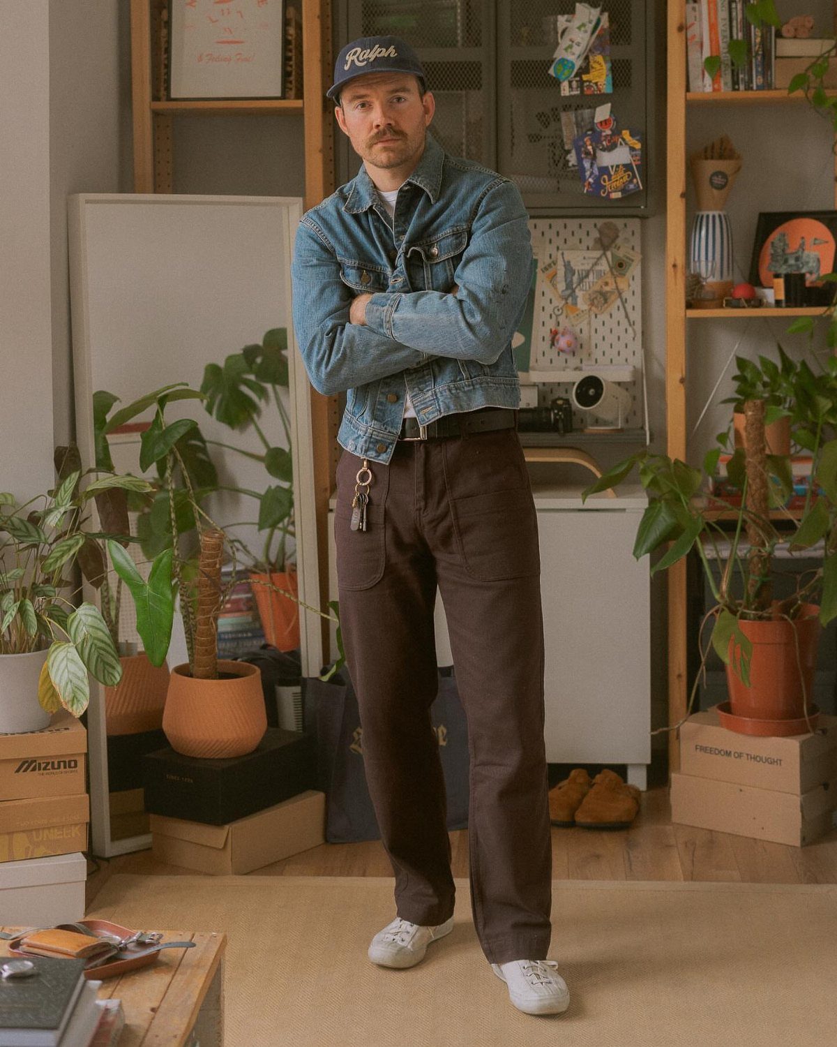 Man in denim jacket and brown trousers standing in a room with many indoor plants.