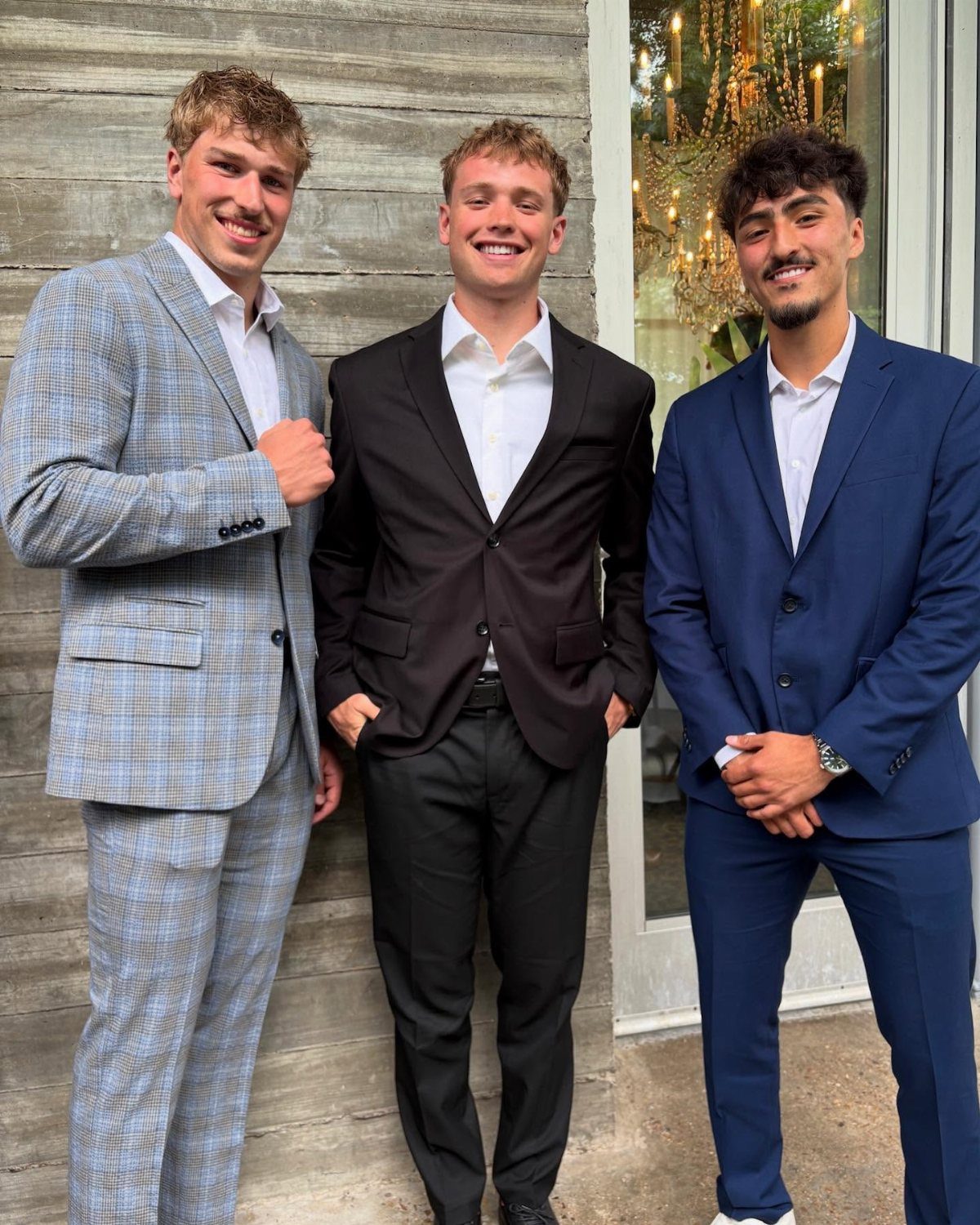 Three young men smiling while wearing stylish suits in light blue plaid, dark brown, and navy blue.