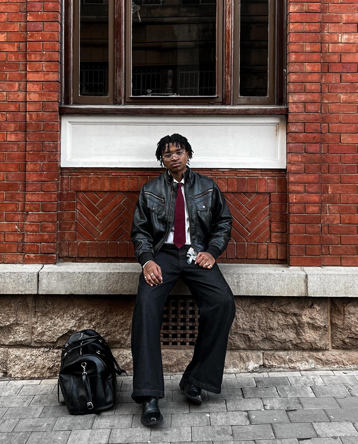 A man in a black leather jacket, red tie, and wide-leg trousers sitting against a brick wall.