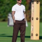 David Beckham wearing a white t-shirt, brown trousers, and a black cap on a grass field.