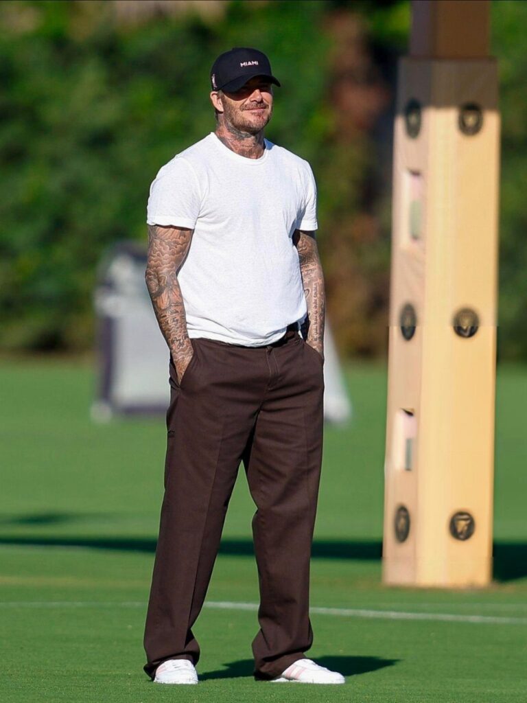 David Beckham wearing a white t-shirt, brown trousers, and a black cap on a grass field.