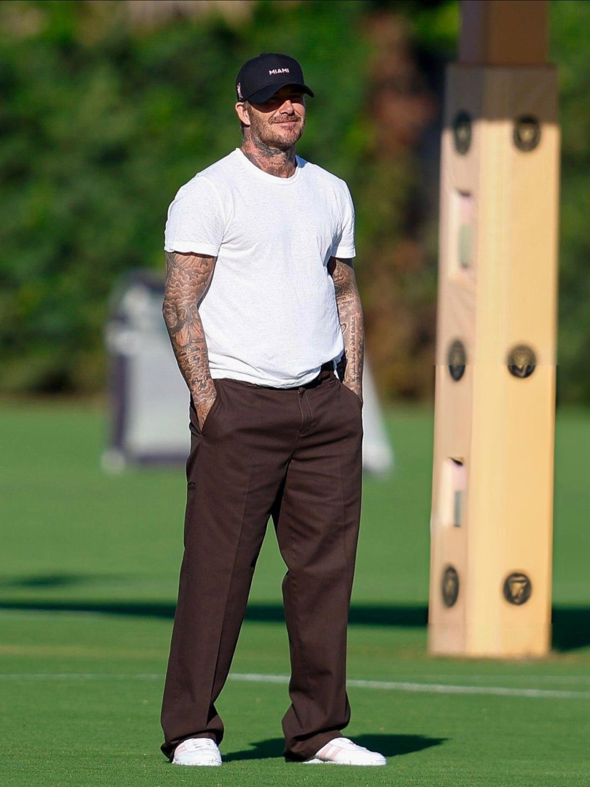 David Beckham wearing a white t-shirt, brown trousers, and a black cap on a grass field.