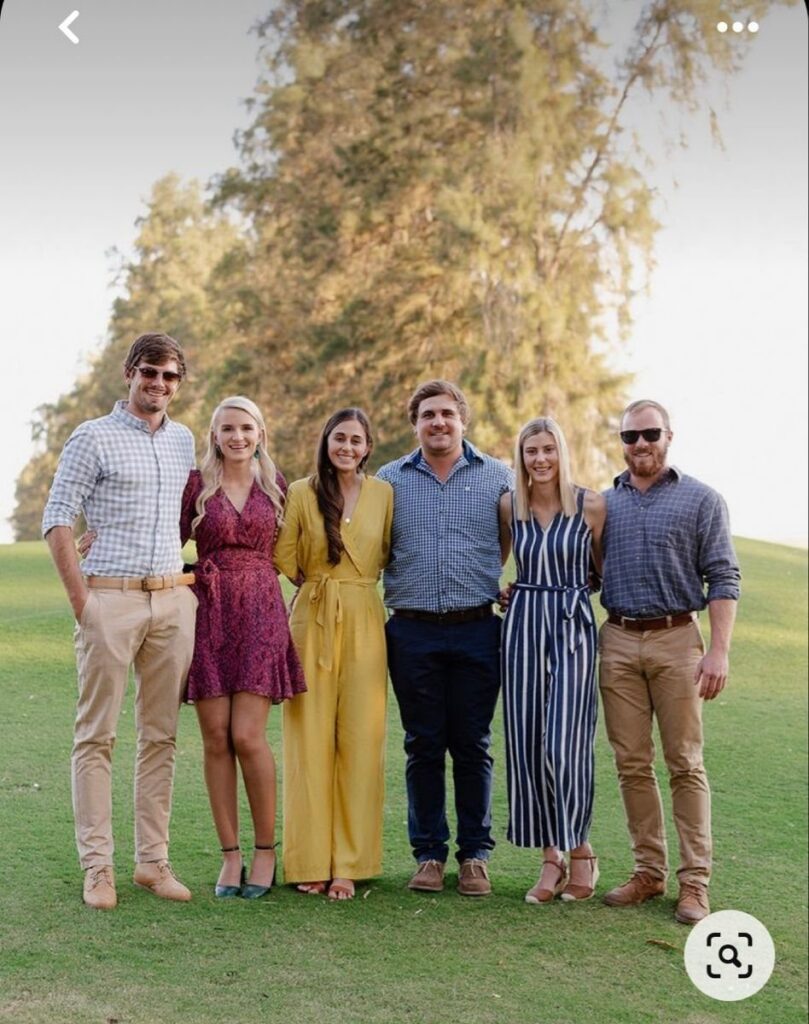 Six young adults posing together in casual attire on a grassy field with trees.