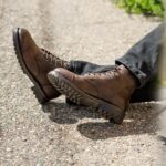 Brown leather lace-up boots worn with dark denim jeans on a concrete surface.