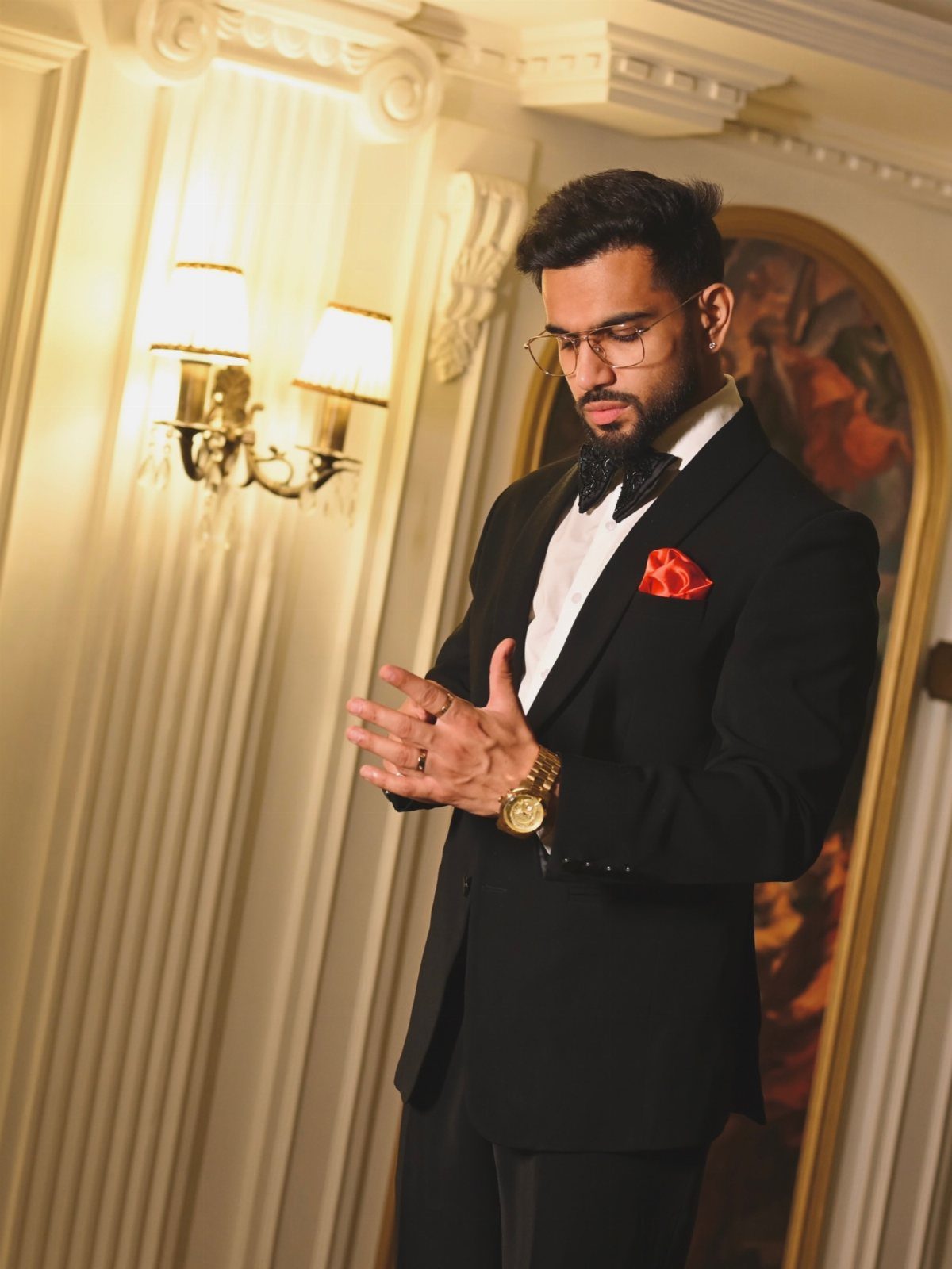 Man in a black tuxedo and white shirt posing in an elegant room with warm lighting.