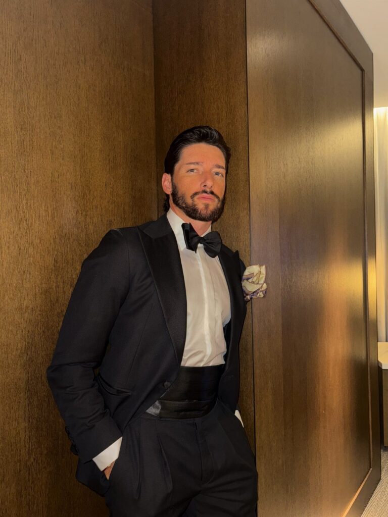 Man in a black tuxedo and bow tie posing against a wooden wall.