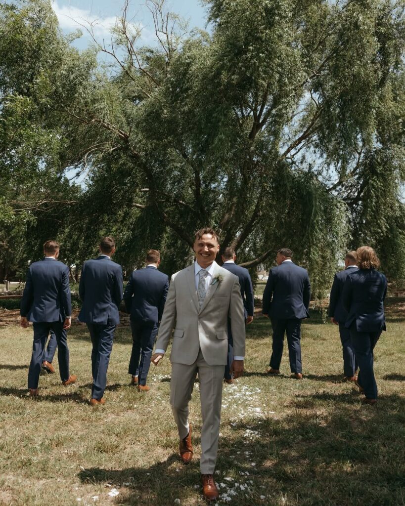 A man in a light grey suit walks toward the camera through a grassy field.