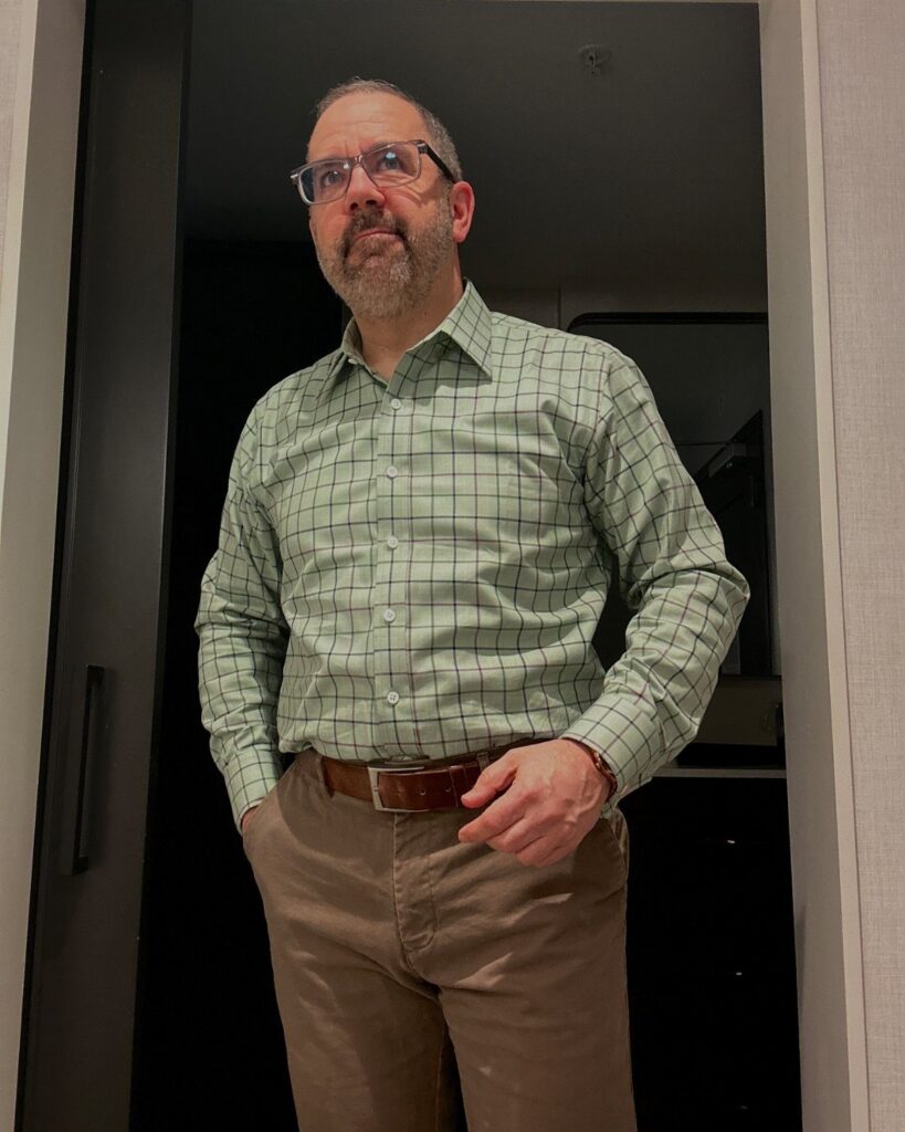 A man wearing a green checkered shirt and brown trousers standing in a doorway.