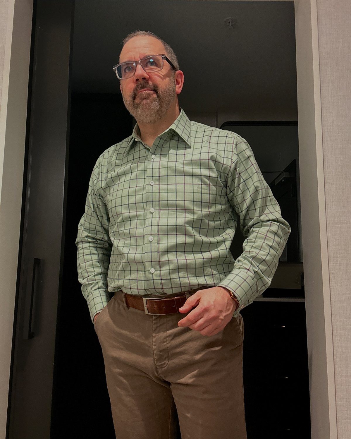A man wearing a green checkered shirt and brown trousers standing in a doorway.