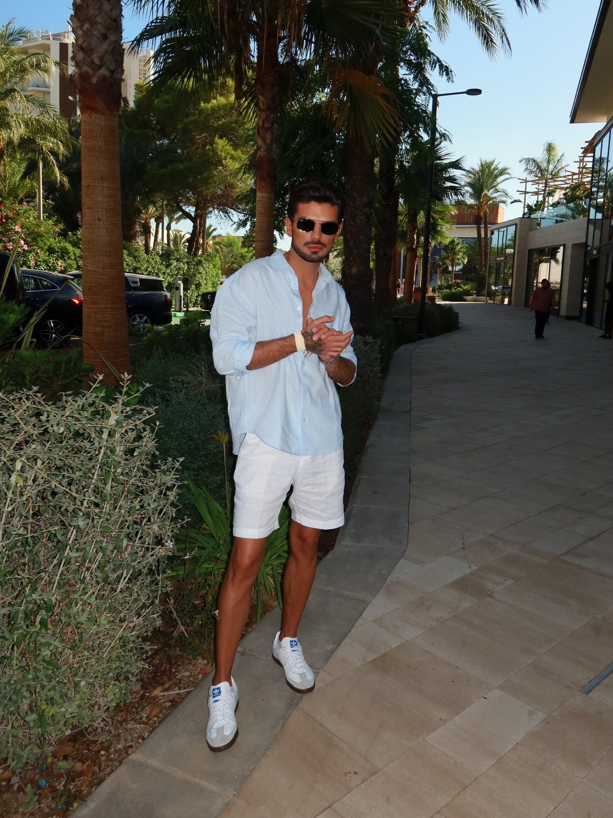Man in a light blue shirt and white shorts standing on a tropical walkway.
