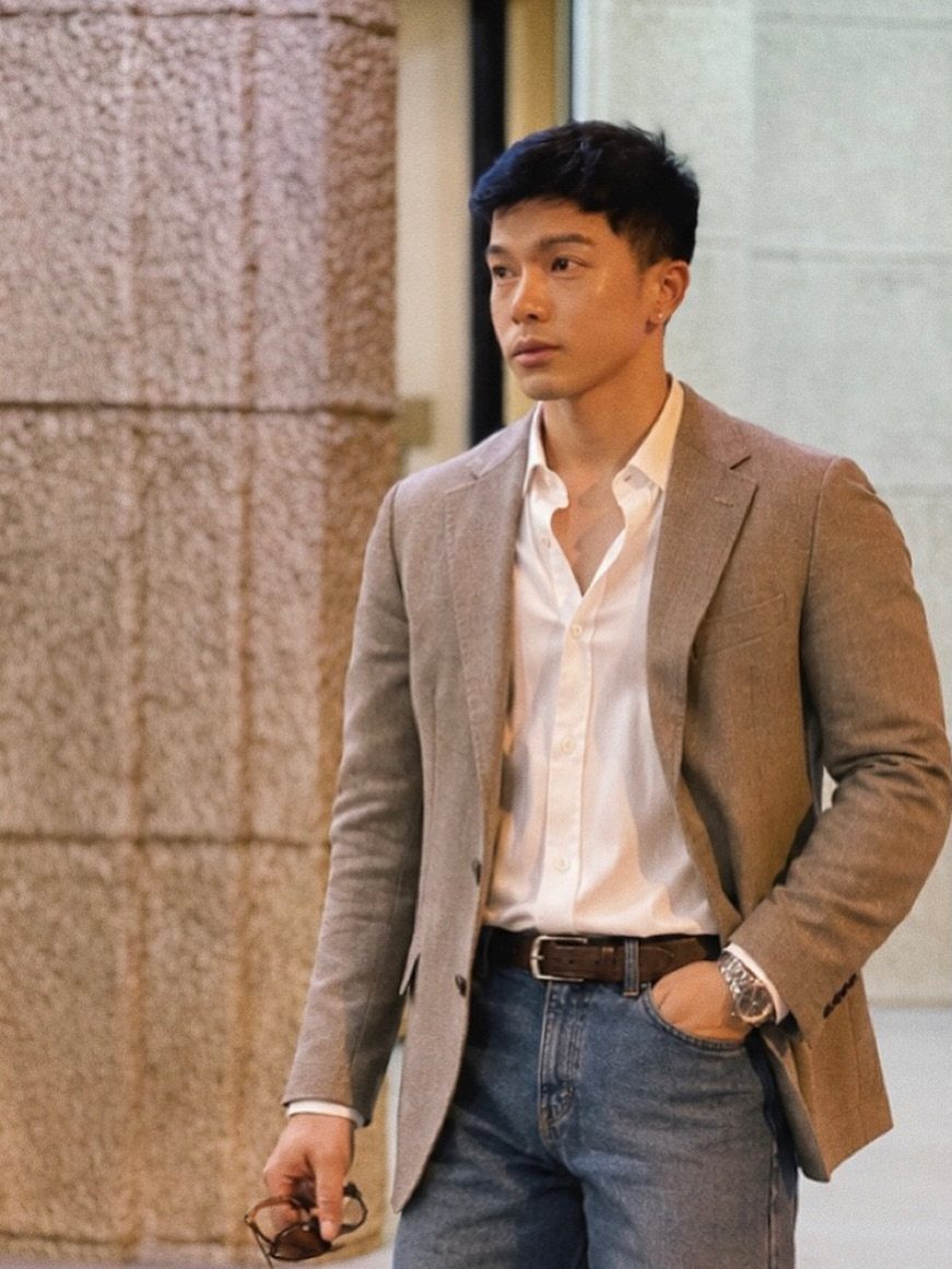 Man wearing a tan blazer, white shirt, and blue jeans standing by a wall.