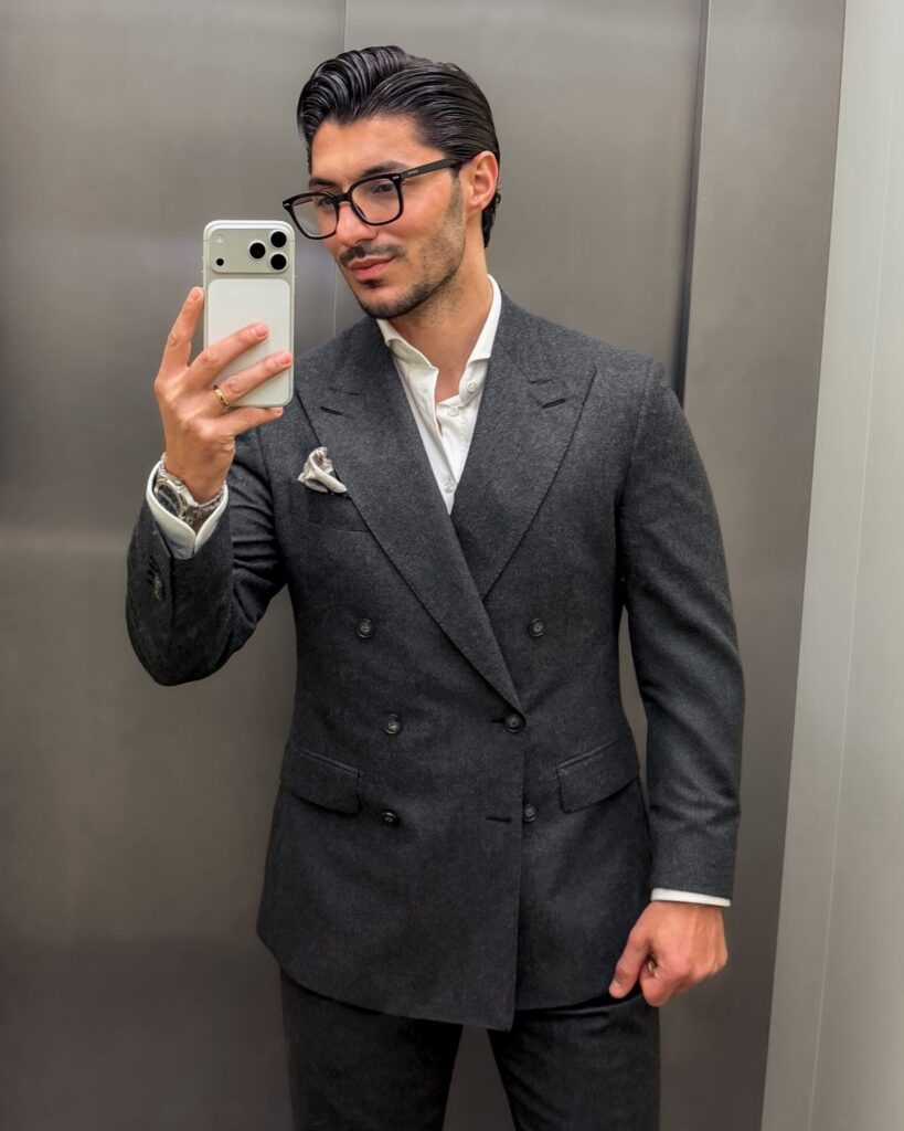 Man in a charcoal double-breasted suit and glasses taking a mirror selfie.