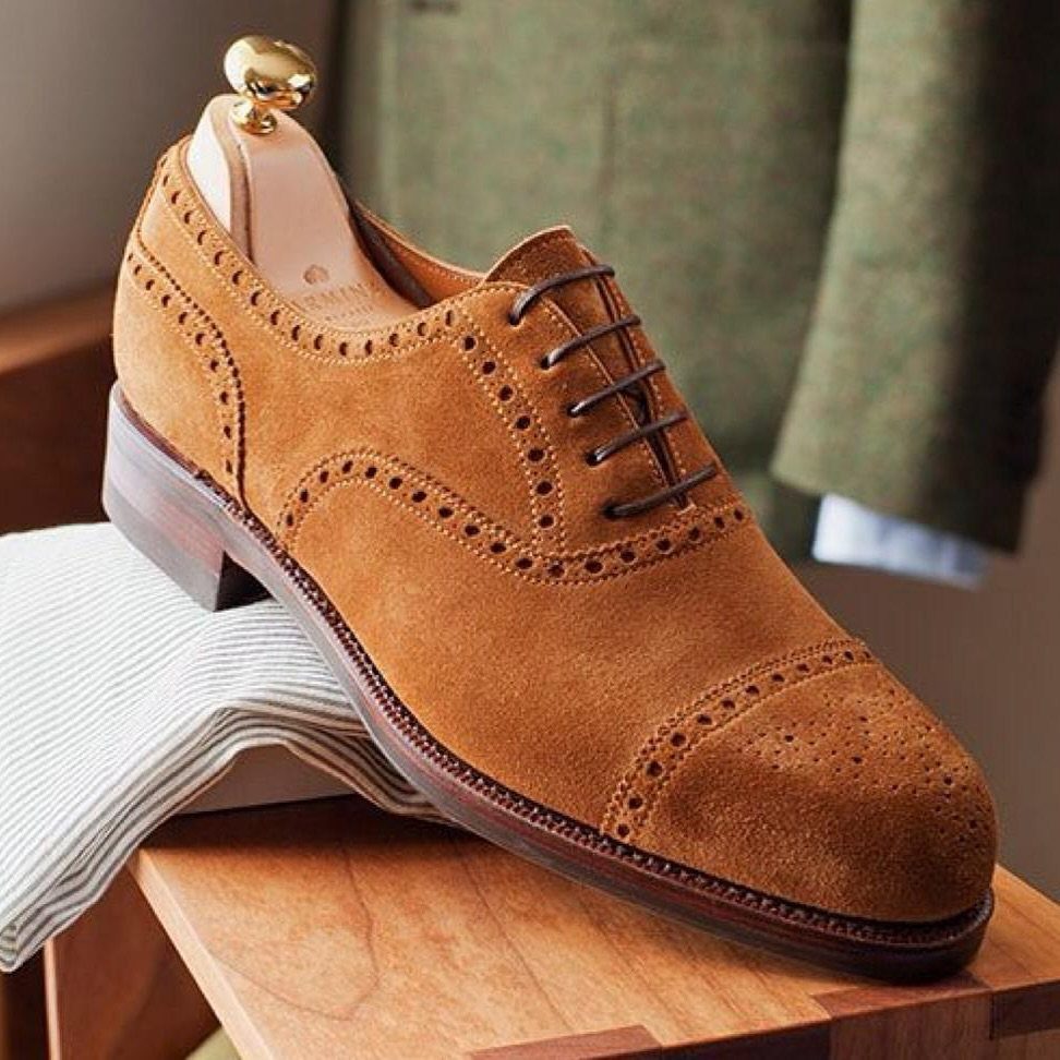 Tan brown suede brogue shoe with decorative perforations resting on a wooden surface.