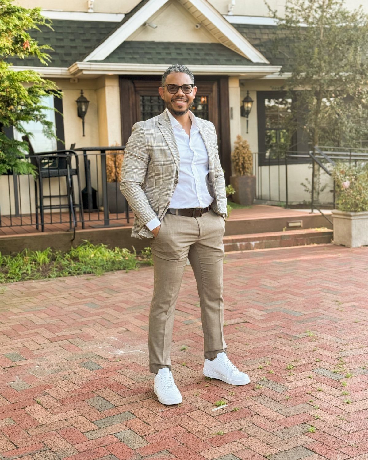 A man wearing a patterned blazer, white shirt, tan pants, and white sneakers standing on a brick patio.