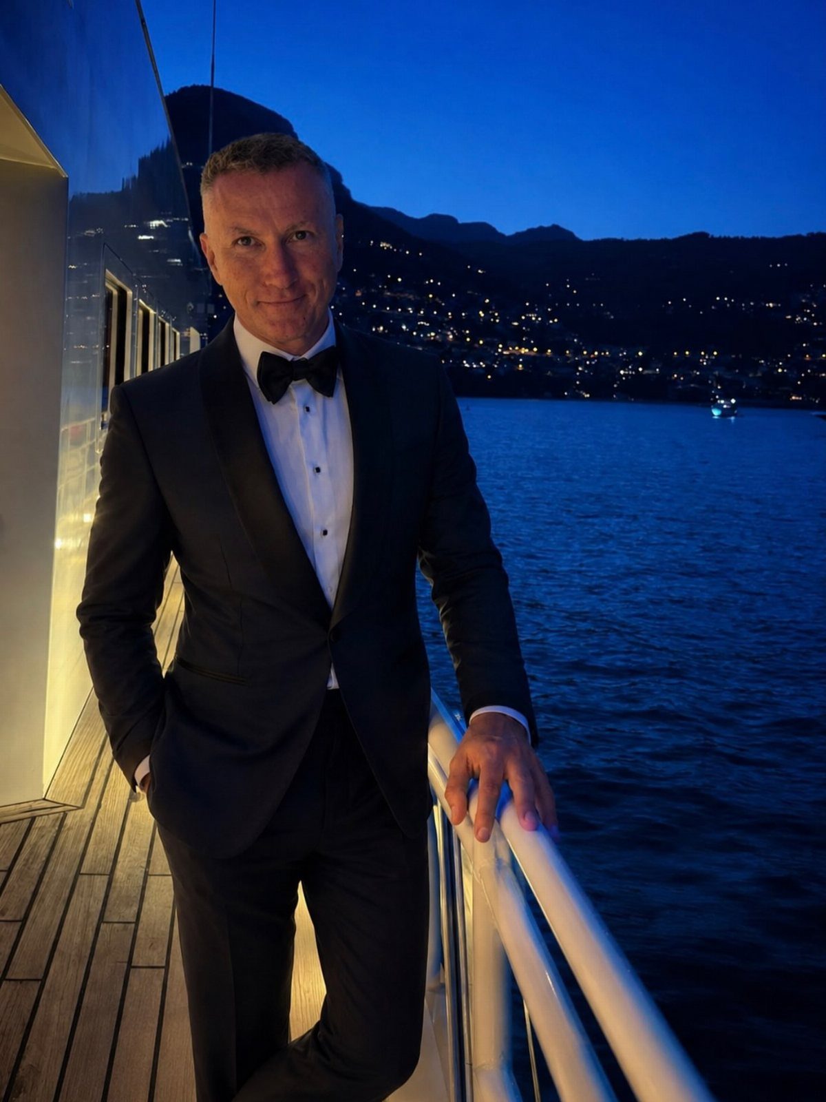 Man wearing a dark navy tuxedo and bow tie posing on a boat at night.