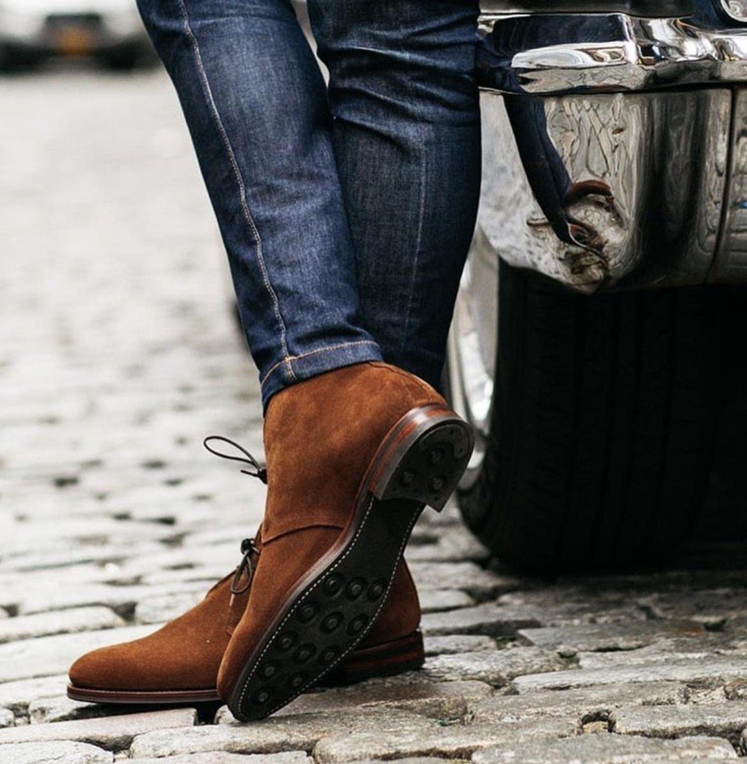Brown suede chukka boots worn with blue denim jeans on a cobblestone street.