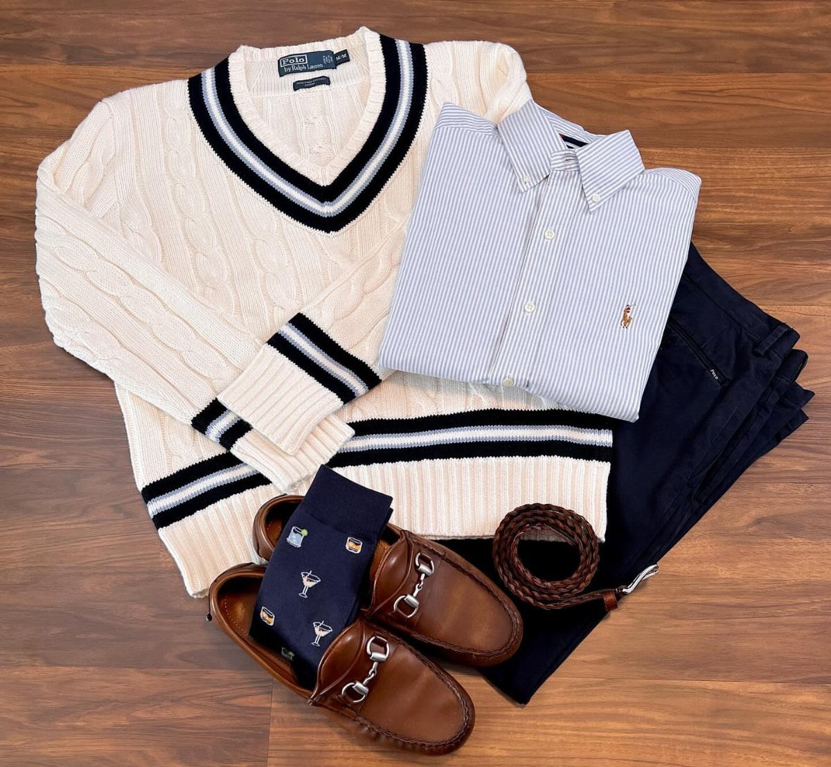 Flat lay of a cream cable-knit sweater, blue striped shirt, navy pants, and brown loafers.