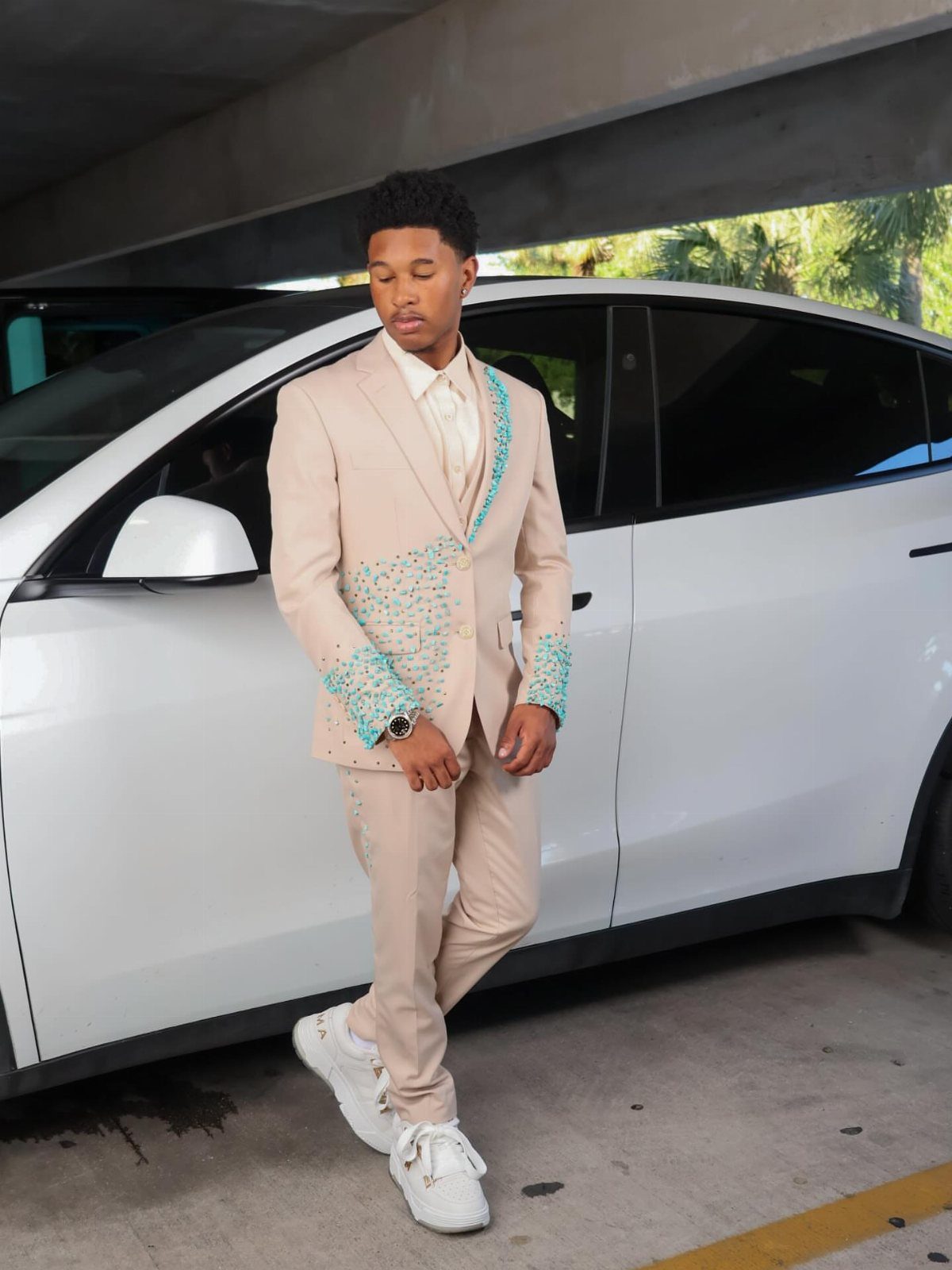A young man in a tan suit with teal sequin details posing in front of a white car.