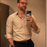 A man in a beige button-down shirt and dark trousers taking a mirror selfie.