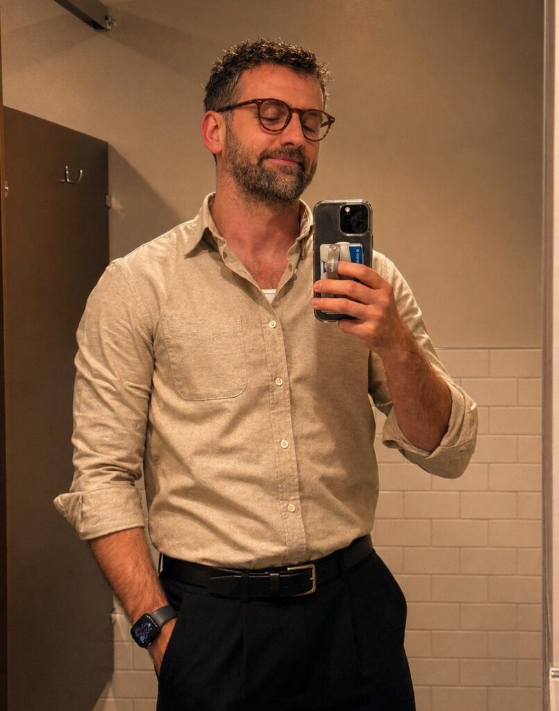 A man in a beige button-down shirt and dark trousers taking a mirror selfie.