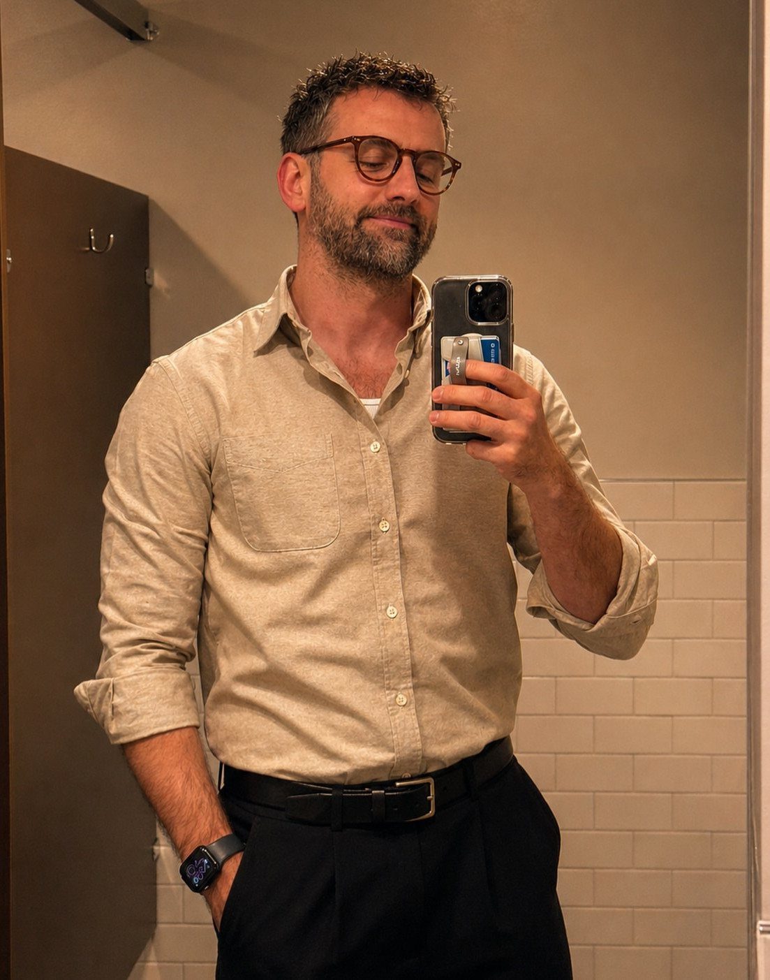 A man in a beige button-down shirt and dark trousers taking a mirror selfie.