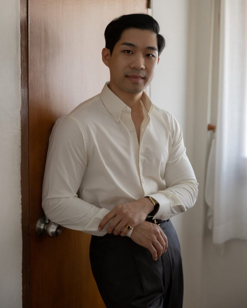 Man in a cream long-sleeved button-down shirt leaning against a wooden door.