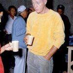A man in a yellow sweater and grey trousers holding a striped mug in a crowd.