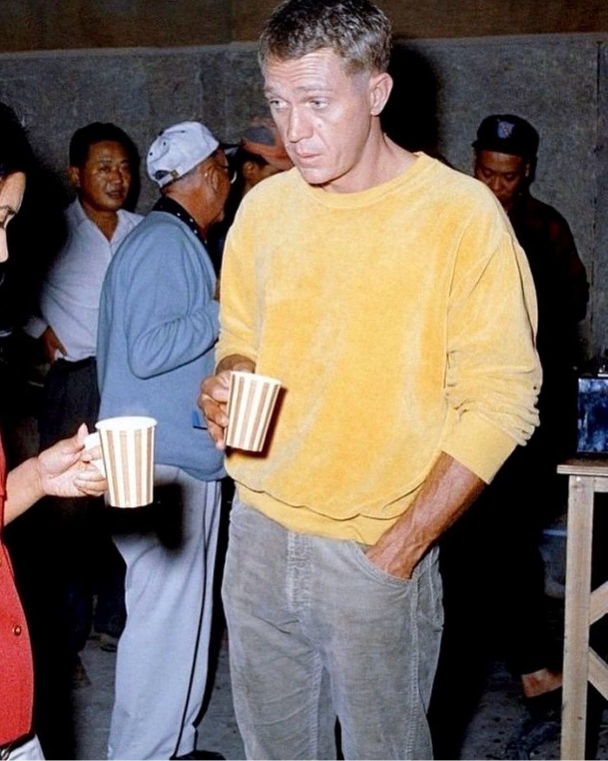 A man in a yellow sweater and grey trousers holding a striped mug in a crowd.