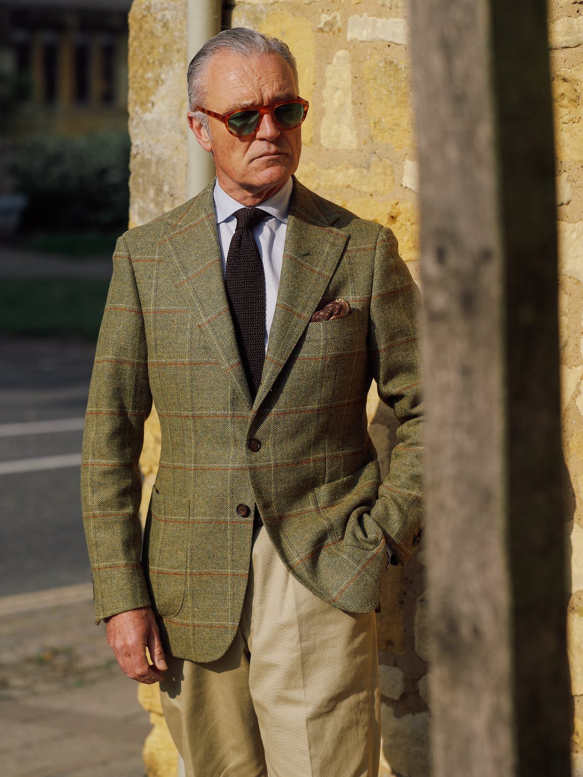 Man wearing a green tweed blazer, patterned tie, and beige trousers standing outdoors.
