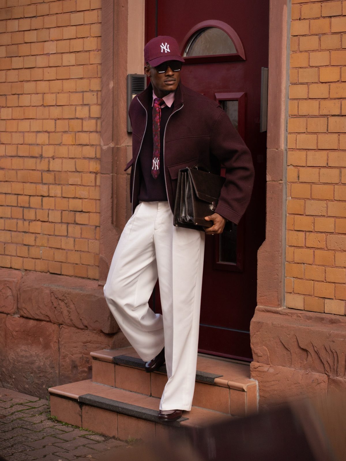 Man in maroon jacket, tie, and white trousers posing on brick steps.